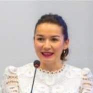 A woman with dark hair tied back, wearing a white lace blouse, speaks into a microphone against a plain light background.