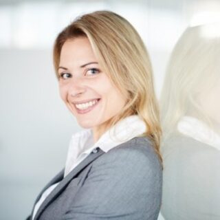 A woman with blonde hair in a gray blazer and white shirt smiles while standing against a glass wall, looking over her shoulder in a brightly lit, modern setting, ready to present on EUDAMED compliance for legacy devices.