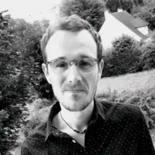 A man with short hair, glasses, and a trimmed beard wears a dark, patterned shirt and a necklace. He stands outside, smiling softly, with trees and houses in the background. The photo is in black and white.