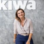 A woman with shoulder-length brown hair, wearing a light blouse and blue pants, smiles while sitting in front of a gray textured wall with the large white letters "kiwa" partly visible behind her, reflecting best practices in professional presentation.