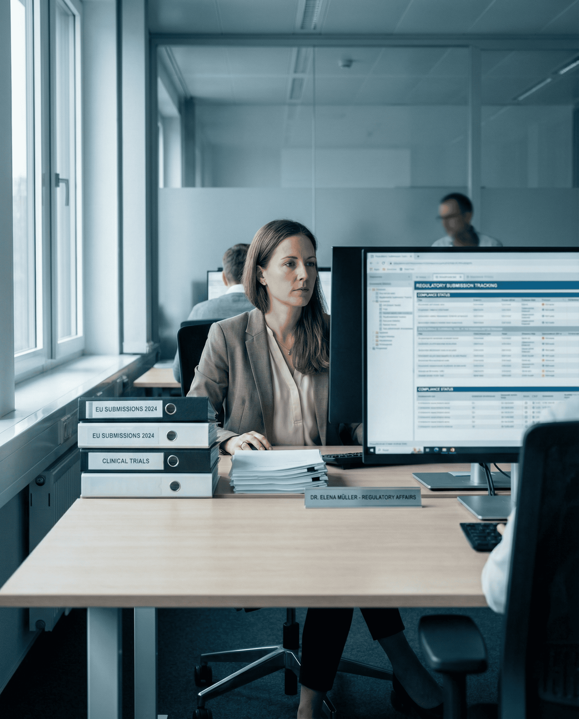 A woman sits at a desk in an office working on a computer. Stacks of binders labeled "EU Submissions 2024" and "Clinical Trials" are on her desk. Other people work in the background.