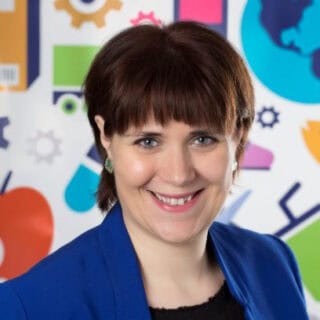 A woman with short brown hair and blue eyes smiles, wearing a blue blazer and earrings. The colorful background features abstract shapes, gears, a globe icon, and symbols reflecting UDI Standards and best practices in the industry.